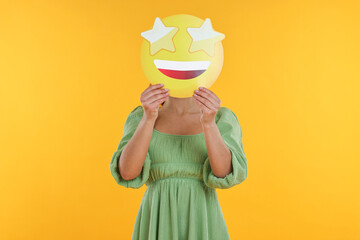 Woman holding emoticon with stars instead of eyes on yellow background