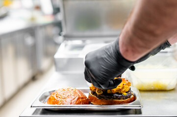 Professional chef in gloves assembling a juicy cheeseburger in a kitchen. Focus on hands with...