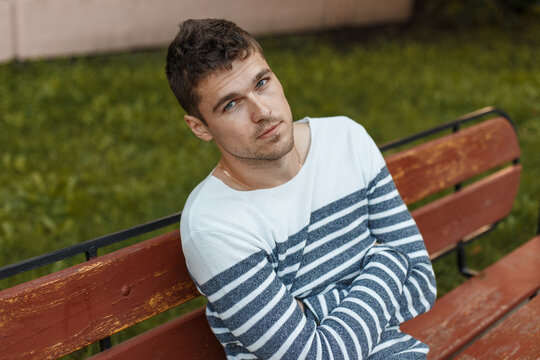 Handsome man model with hairstyle in a sweater sits on a bench in the park