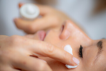 Cosmetician delicately applies cream on woman's face, known for its moisturizing and rejuvenating...