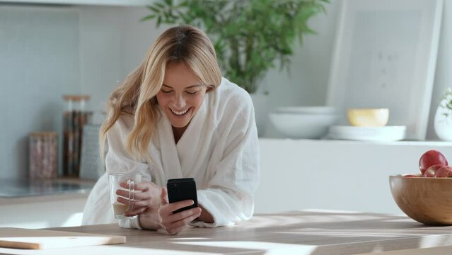 Video of smiling young woman enjoying a cup of coffee while using her mobile phone in the living room at home.