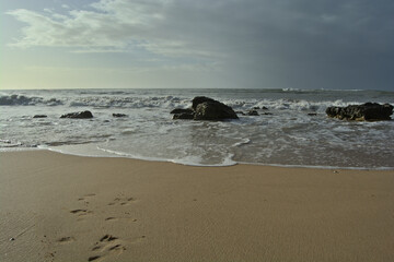 Photo of the shoreline on a Portugese beach. Footprints in the sand and water crashing on the rocks.
