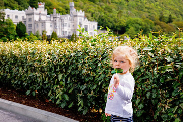 Cute toddler girl with Irish cloverleaf lollipop with Kylemore Abbey on background. Happy healthy child on flower meadow eating unhealthy sweets. Family and small children vacations in Ireland