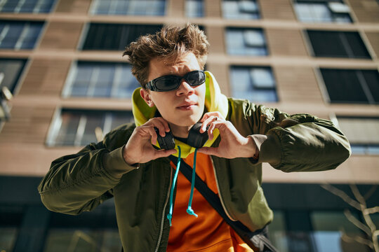 Portrait of young attractive guy, in trendy vibrant outfit and stylish, retro sunglasses posing against urban cityscapes. Concept of street fashion, style, modern lifestyle, gen Z, self-expression.