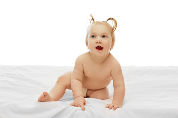 Little cute baby girl, child in diaper, with two ponytails sitting on white bedsheets with curious playful expression over white background. Concept of childhood, care, health, well-being, parenthood