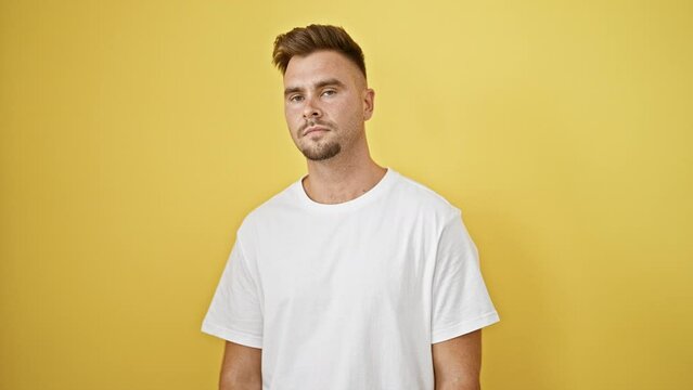 Handsome young adult hispanic man with a beard standing against a solid yellow background in a white shirt, looking to the side.
