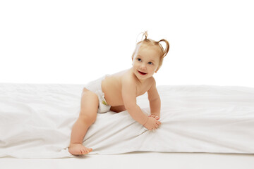 Little cute baby girl, child in diaper, with two ponytails playing on white bedsheets with happy expression against white background. Concept of childhood, care, health, well-being, parenthood