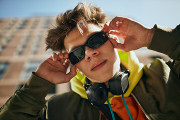 Creative portrait of young attractive guy, in trendy vibrant outfit and stylish, retro sunglasses posing against cityscapes. Concept of street fashion, modern lifestyle, gen Z, self-expression.