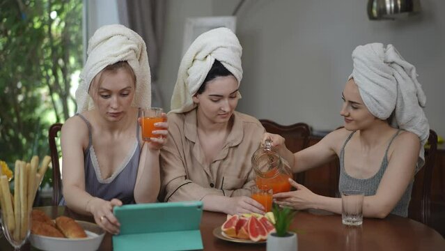 Three girls with towels on their heads pour orange juice from a glass jug into glasses, while sitting at a table in the living room in the morning