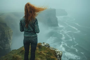 Foto op Plexiglas A solitary woman in a denim jacket stands facing the ocean, her hair blowing in the wind atop a mist-covered cliff © Larisa AI