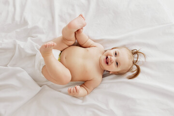 Happy, smiling, little redhead girl, toddler in diaper lying on belly on bed, laughing, playing on white bedsheets. Happiness, fun and joy. Concept of childhood, care, health, well-being, parenthood