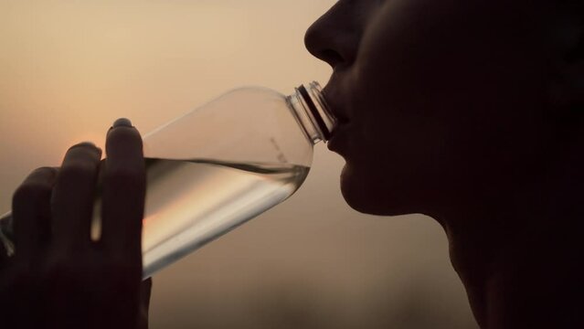 Woman drinking water after training. Fitness thirsty girl drinks water during workout pause, after a grueling run of a long sea ocean beach at sunset. Endurance sport, relaxing, morning run concept.