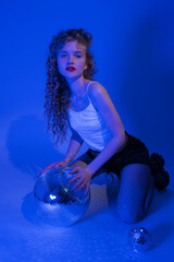 Young woman with disco ball is posing in music style while partying in nightclub. Sensuality woman kneeling in blue neon colored lights. Beautiful woman in white crop top, black leather shorts, boots