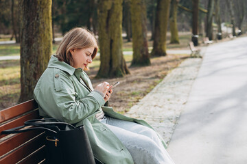 Fototapeta na wymiar 30s cheerful woman tourist dressed in casual look holding smartphone gadget in hand for communicate on the city street. Happy woman chatting on cell smartphone and relaxing on the bench outdoors