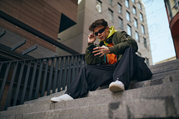 In heart of city's rhythm. Cool attitude young man sitting using phone on stairs against skyline,...