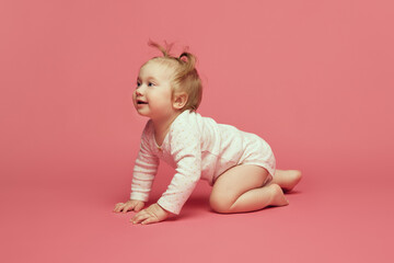 Smiling happy little baby girl, toddler in comfortable onesie crawling, playing on pink background. Joyful time. Concept of childhood, care, health, well-being, parenthood