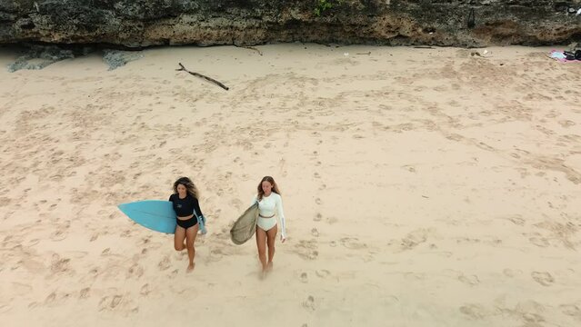 Aerial view of two young diverse women in black and white bikinis walking along the beach towards the ocean with surfboards under their arms. Surfing in Bali. Women in extreme sports.