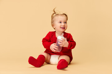 Little baby girl, child with ponytails in red dress and socks holding bottle, smiling, sitting on beige background. Concept of childhood, care, health, well-being, parenthood
