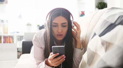 Young beautiful woman headphones holds smartphone in hand using at home costs about sofa. Verifies internal information its financial advertising freedom makes money level transfer contractor concept