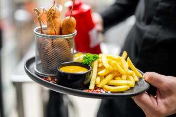 Close-up of a server holding a round tray with crispy French fries, yellow dipping sauce, and three corn dogs. A piece of broccoli garnishes the food. Background includes a red ketchup bottle
