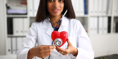 Beautiful black smiling female doctor hold in arms red toy heart closeup. Cardio therapeutist student education CPR 911 life save physician make cardiac physical pulse rate measure arrhythmia