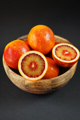 Lots of cut and whole blood oranges in a wooden bowl on a dark background