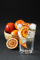 Glass with water, ice and citrus next to blood oranges on a dark background