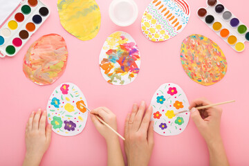 Mother and baby girl hands holding paintbrush and painting colorful eggs on paper with watercolor on pink table background. Closeup. Child making easter decoration. Point of view shot. Top down view.