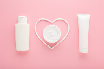 White heart shape with cream jar, bottle and tube on light pink table background. Pastel color. Care about female face, hands, legs and body skin. Closeup. Top down view. - 783033146
