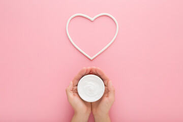 Young adult woman hands holding white cream jar. White heart shape on light pink table background. Pastel color. Care about clean and soft female body skin. Closeup. Point of view shot. Top down view. - 783033156
