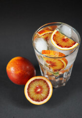 Glass with water, ice and citrus next to blood oranges on a dark background