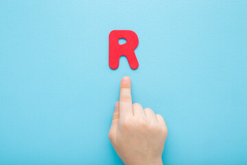 Baby boy hand finger pointing to red R letter on light blue table background. Pastel color. Time to...