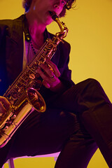 Cropped portrait of solo player in black suit playing sax in neon light against vibrant yellow...
