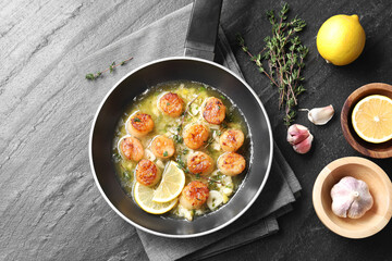 Delicious scallops with sauce in frying pan and ingredients on dark gray textured table, flat lay