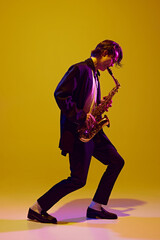 Obraz premium Young virtuoso in classical black suit playing saxophone in neon light against vibrant yellow studio background. Concept of music and art, hobby, concerts and festivals, modern culture. Ad