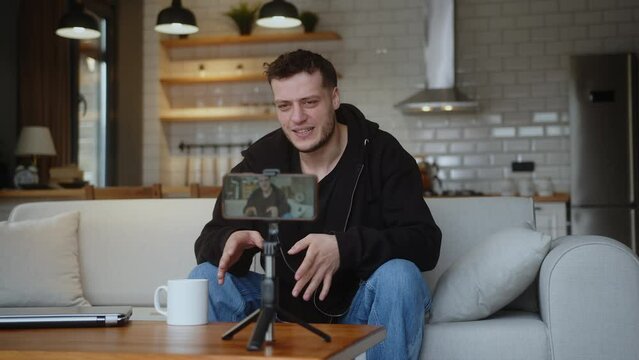 Young influencer vlogger man sit on the couch recording video blog on mobile camera tripod talk for personal media channel, streaming live session blogging broadcast at home.