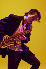Portrait of young attractive man in dark attire performing solo with sax in neon light against vibrant yellow studio background. Concept of music and art, hobby, concerts and festivals, modern culture