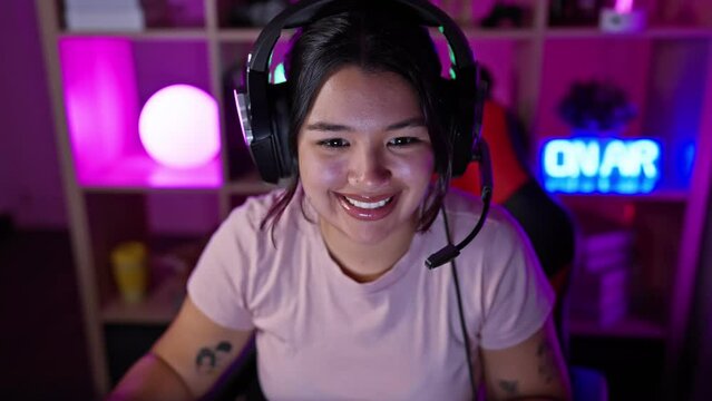 A joyful hispanic woman with headphones in a vibrant gaming room smiling at the camera indoors at night.