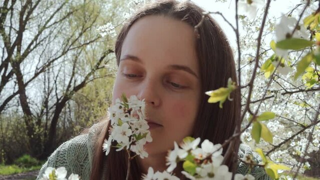 Caucasian young adult woman sneezing while posing among blooming cherry trees in flowering garden using napkin during springtime allergy