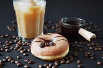 Caramel donut with coffee decor next to a glass of frappuccino with ice and a glass scoop of coffee on a dark background