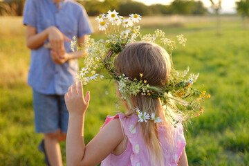 boy holding doll woven from grass in hand, happy girl in floral crown, wreath holding chamomile flower in hand on green sunlit meadow, natural world, happy childhood In heart nature