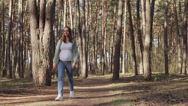 Satisfied extremely happy cheerful pregnant woman resting in forest enjoying beautiful nature and pine-tree wood walking in park breathing fresh air wearing casual clothing