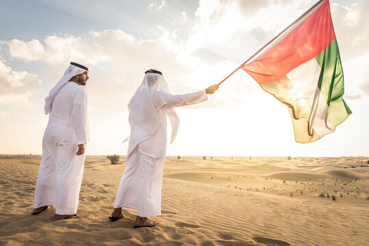 Two arab men wearing traditional emirati clothing in the desert of Dubai - Middle-eastern adult males portrait holding emirate flag to celebrate national day
