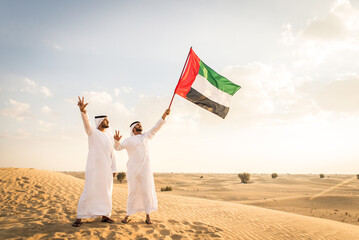 Two arab men wearing traditional emirati clothing in the desert of Dubai - Middle-eastern adult males portrait holding emirate flag to celebrate national day - obrazy, fototapety, plakaty