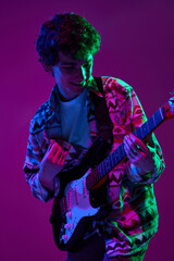 Fototapeta premium Young artistic guitarist in retro outfit performing in pink-purple stage neon light against gradient studio background. Concept of music and art, hobby, concerts and festivals, modern culture. Ad
