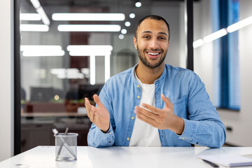Photo of a smiling young hispanic man in a denim shirt sitting at a desk in a happy office and...
