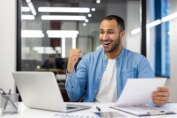 Happy young muslim man sitting in modern office at work desk, looking at laptop screen, holding documents and enjoying success, showing victory gesture with hand