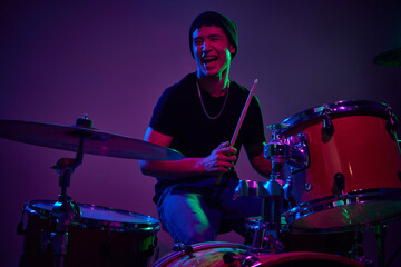 Fototapeta na wymiar Joyful contemporary drummer playing in pink-purple stage lighting against gradient studio background. Concept of music and art, hobby, concerts and festivals, modern culture. Ad