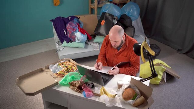 Millennial man anxious desperate worker annoyed work crumpling sheet of paper throw away working in messy home living room with scattered stuff. Mess, disorder and interior concept