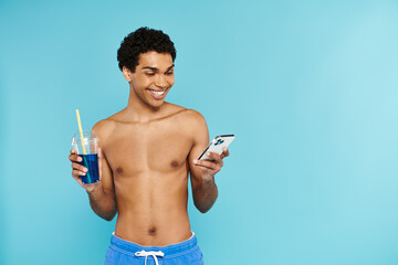 joyful african american man in swimming trunks looking at his phone and holding cocktail in hand